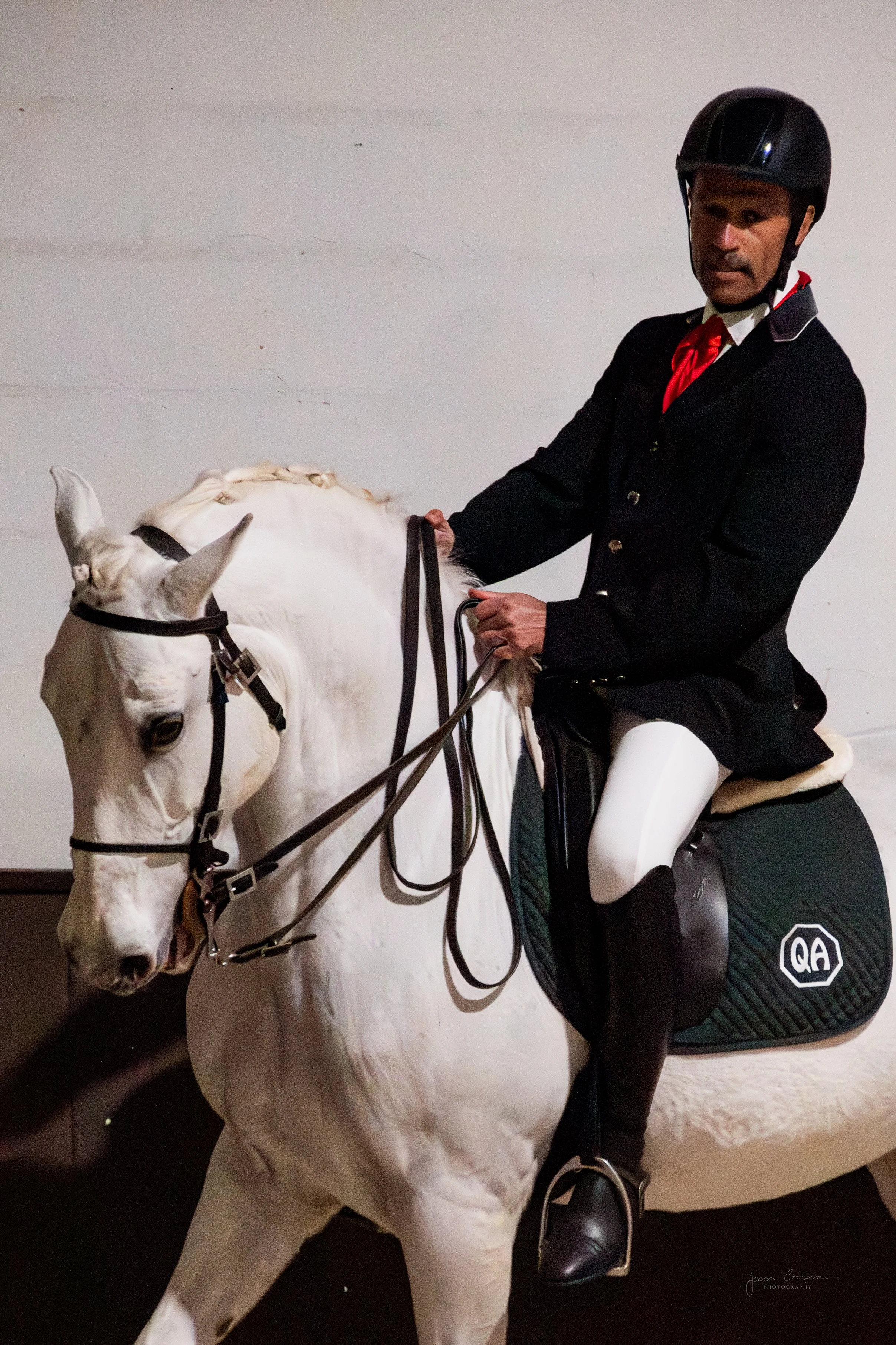 A man dressed in equestrian riding attire sitting on a white horse indoors, wearing a black riding helmet and a dark jacket.