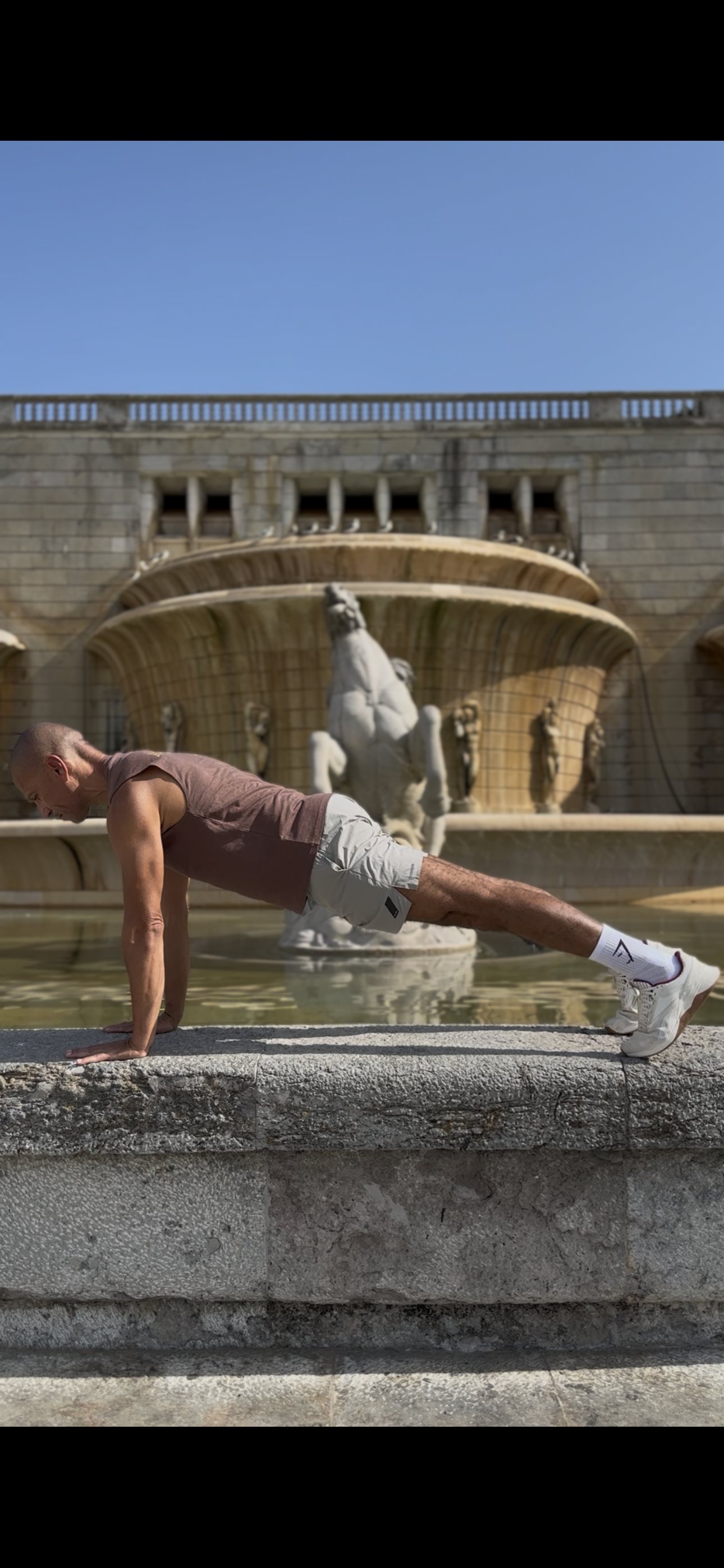 A man doing a plank exercise on a stone edge in front of a fountain with a horse sculpture and statues in a public square.