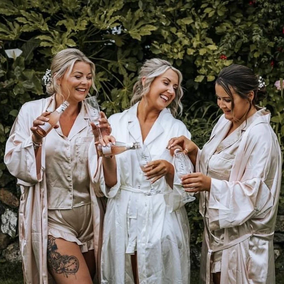 Three women in satin pajamas sharing drinks and laughing outdoors surrounded by greenery.