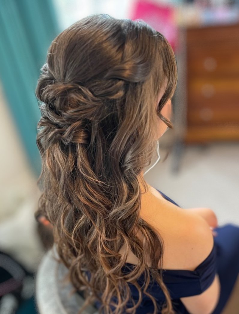 A woman with long, wavy brown hair styled in a side braid and loose curls, sitting indoors with a blurred background.