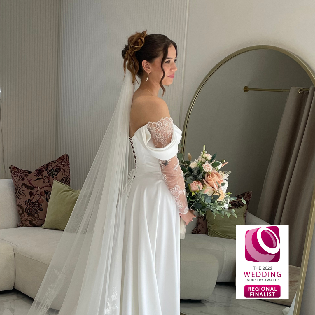Bride in a white wedding gown with off-the-shoulder lace sleeves, holding a bouquet of pink and white roses, standing in front of a mirror in a modern room with beige walls and colorful pillows on a sofa. There is a logo for the 2026 Wedding Industry