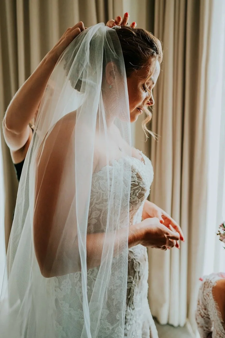 A bride in a wedding dress with a lace bodice is having her veil adjusted by someone behind her. She has curly hair and is standing near a window with curtains, with sunlight illuminating her face.