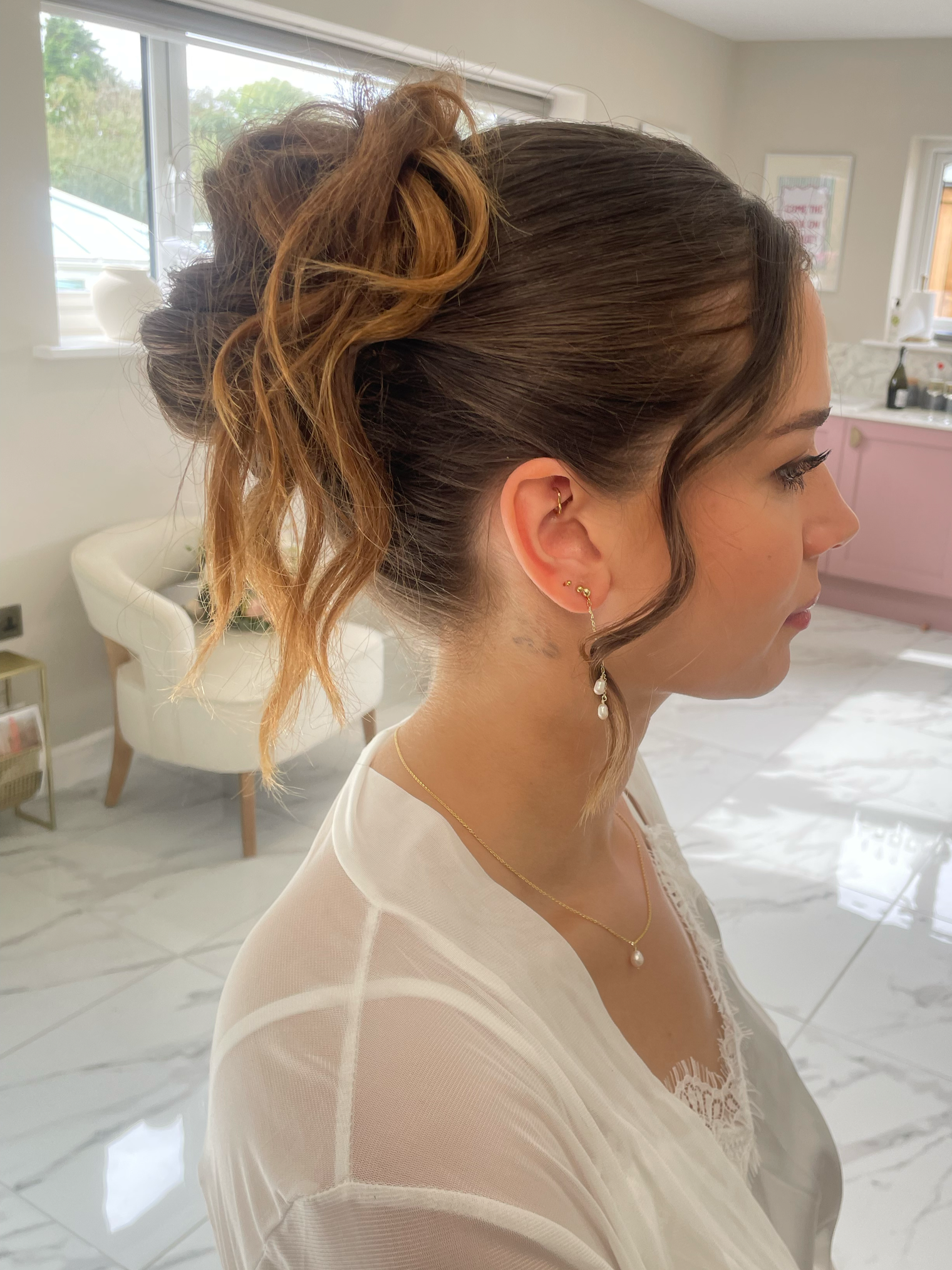 A side profile of a woman with styled hair in an updo, wearing pearl jewelry and a white sheer top, in a bright room with white marble floors and pink cabinetry in the background.
