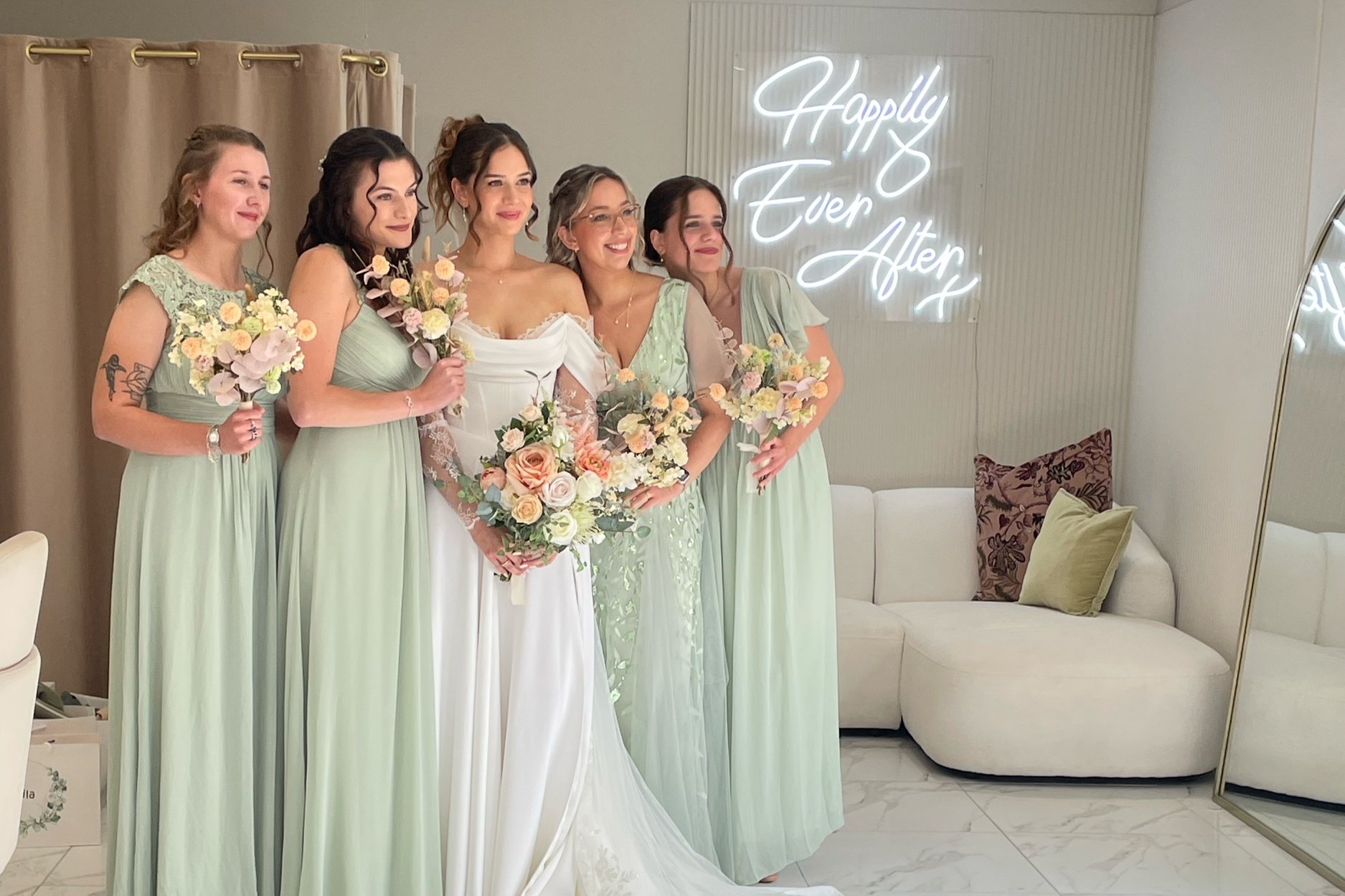 Bride and five bridesmaids posing for a photo in a room with a white sofa, decorative pillows, and a neon sign that reads 'Happily Ever After'.