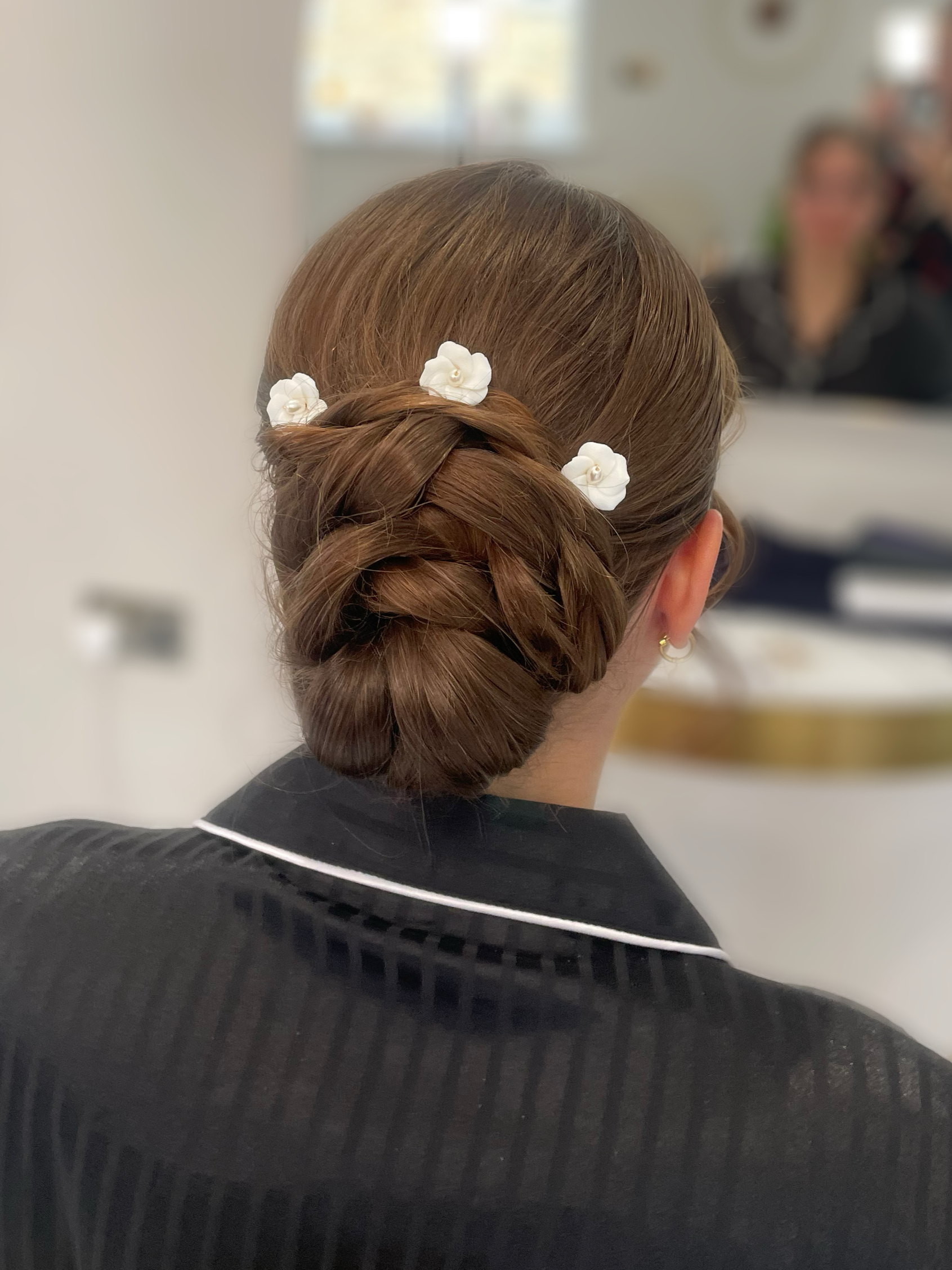 Back view of a woman with brown hair styled in an elegant updo decorated with white floral hair accessories, wearing a black top with white piping, in an indoor setting.