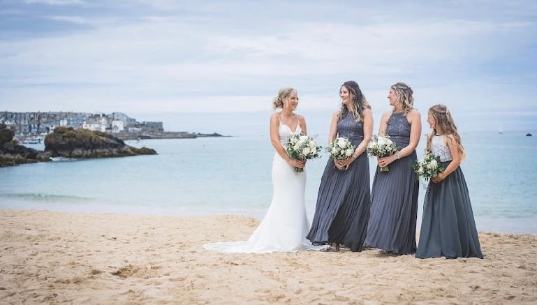 A bride in a white wedding dress talking with three bridesmaids in dark gray dresses on a beach with the ocean and cloudy sky in the background.