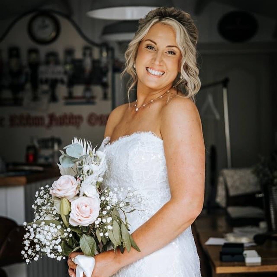 A smiling woman in a white wedding dress holding a bouquet of flowers inside a room.