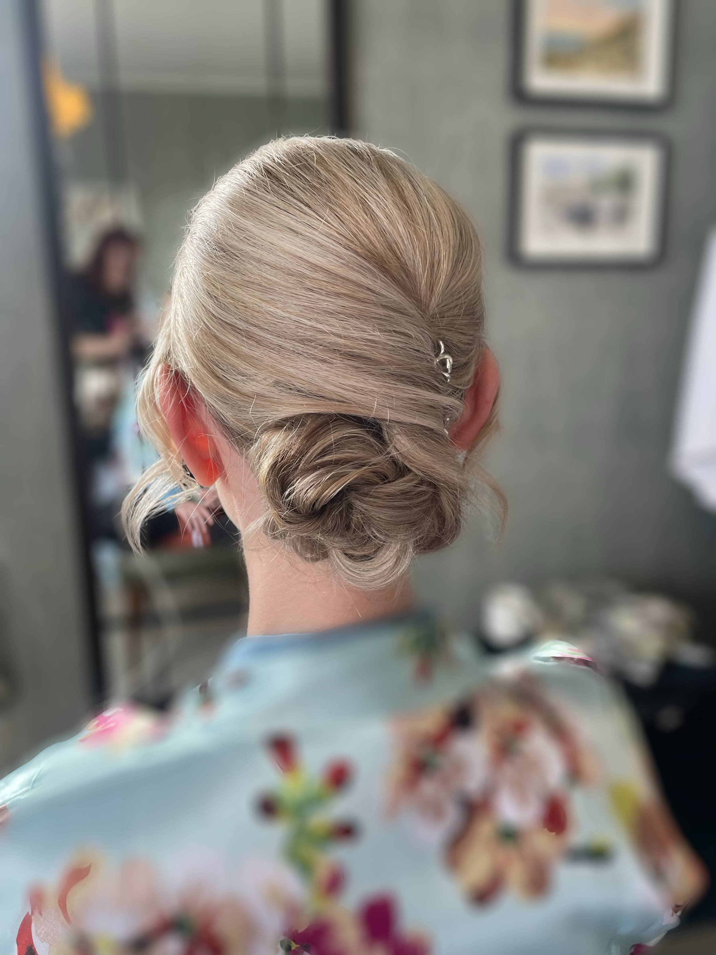 A woman with blonde hair styled in an elegant low bun with a silver hair clip, wearing a floral robe, and facing a mirror in a salon.