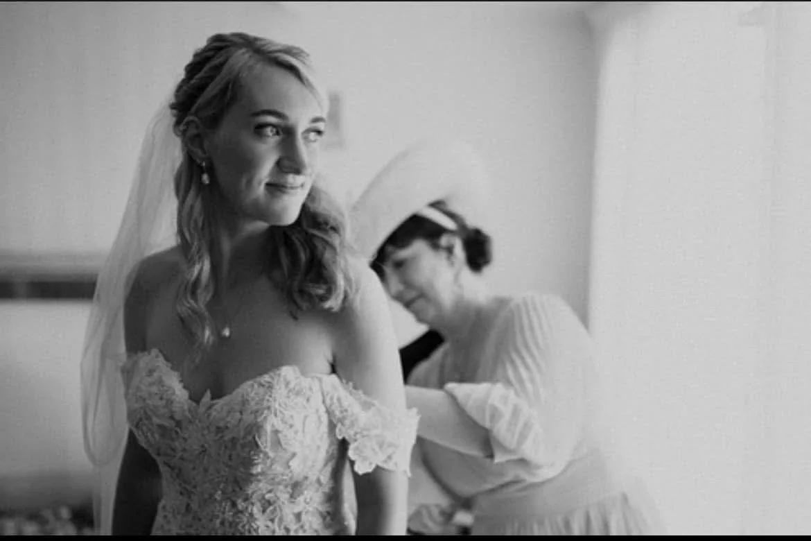 Black and white photo of a bride in a wedding dress with a veil, looking to the side, while a woman in the background, likely her mother or a stylist, is adjusting her dress.