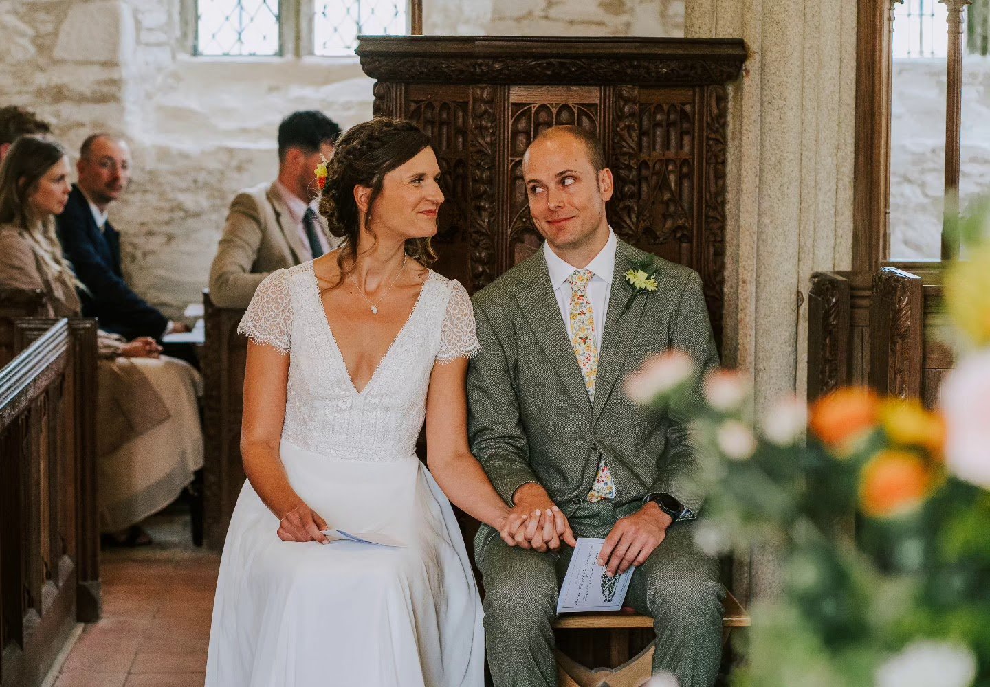 A wedding ceremony with a bride and groom sitting hand in hand in a church or chapel, surrounded by guests, with the bride wearing a white dress and the groom in a suit with a floral tie.