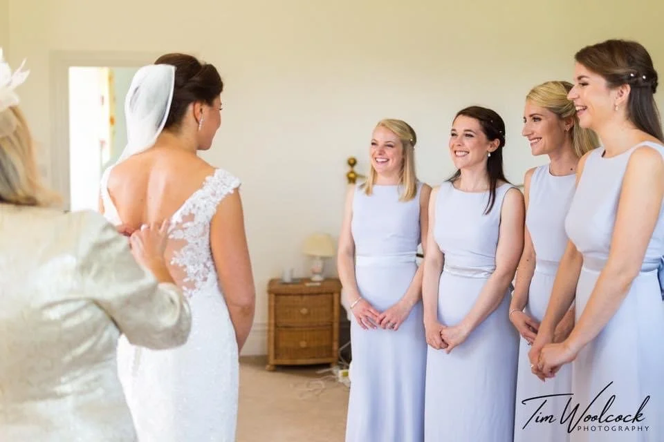 A bride in a lace wedding dress and veil is seeing her bridesmaids, who are dressed in matching light blue dresses, smiling and standing in a line in a bedroom.