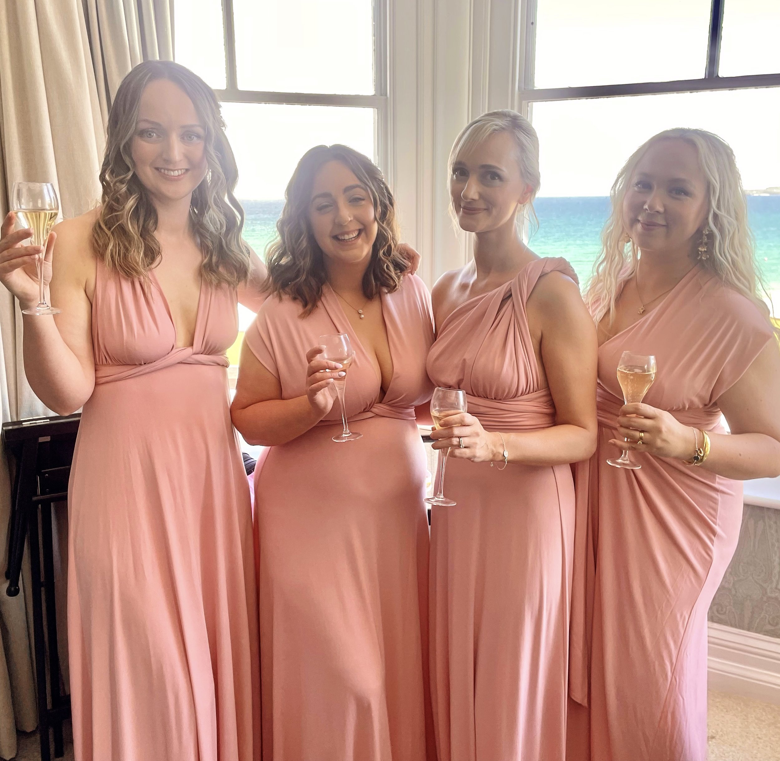 Four women in matching pink dresses holding glasses of champagne, standing indoors near large windows with a view of the ocean in the background.