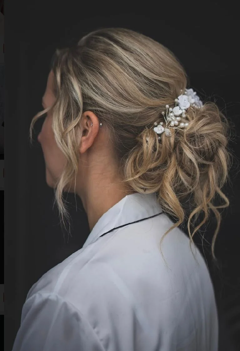 A woman with styled blonde hair in an updo, decorated with white floral hair accessories and pearls, wearing a white garment with a black trim.