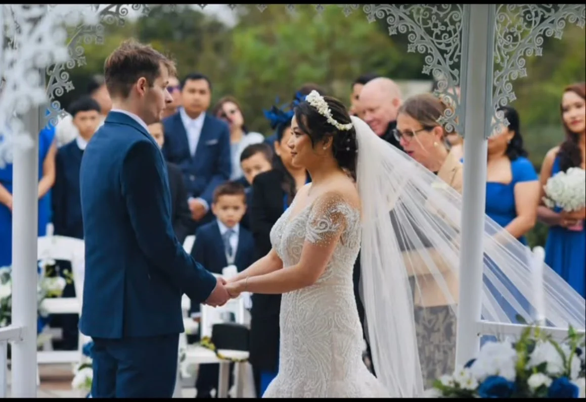 A bride and groom hold hands during an outdoor wedding ceremony with guests in the background.