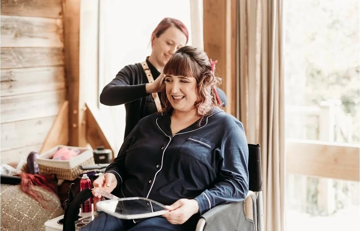 Woman in pajamas sitting in a wheelchair smiling at a tablet, getting her hair styled by a woman standing behind her in a cozy room with large windows and wooden walls.