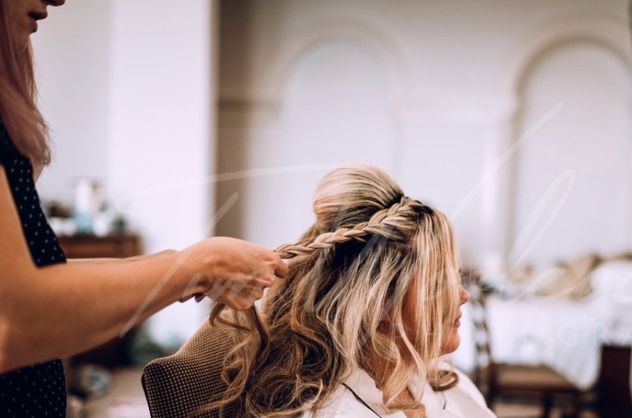 A woman styles another woman's hair in a salon, twisting a section of blonde hair into a braid.
