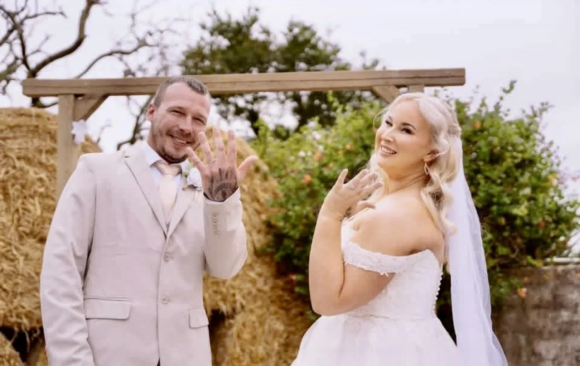 A happy couple in wedding attire showing their wedding rings outdoors. The groom is wearing a light-colored suit and smiling, while the bride is in a white wedding dress with off-the-shoulder sleeves and a veil, also smiling. They are standing in fro