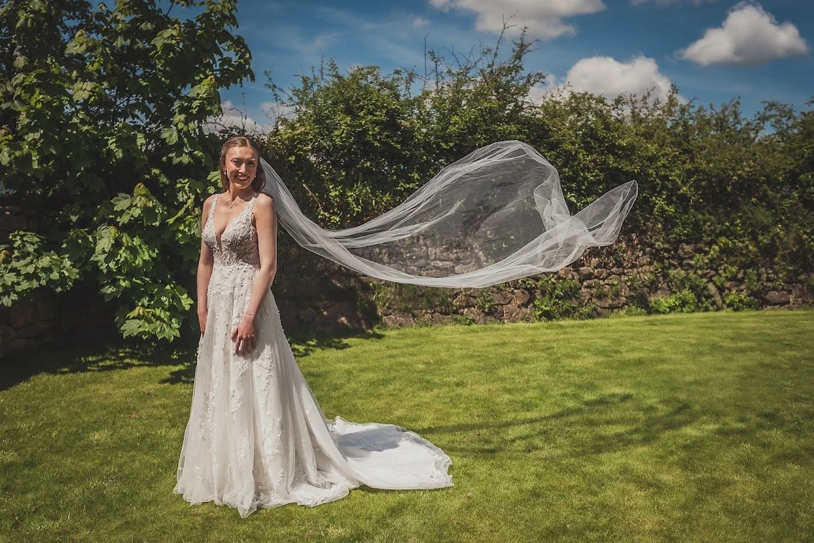 A bride in a wedding dress outdoors on a sunny day, with her veil flowing in the breeze, standing on a green lawn with trees and a stone wall in the background.