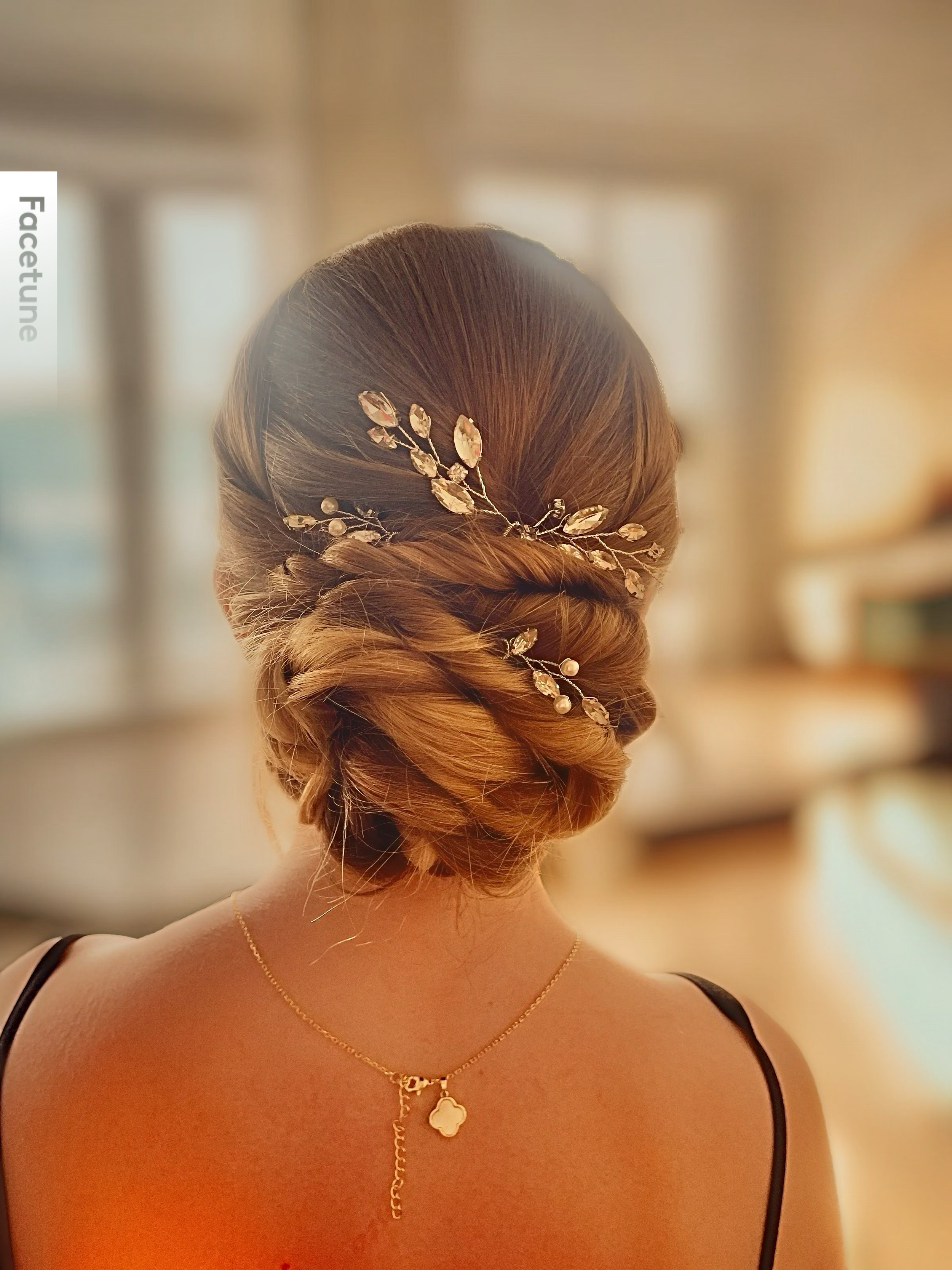 Back view of a woman with styled brown hair adorned with a decorative hairpiece featuring leaves and beads, wearing a gold necklace with a four-leaf clover pendant, and a black spaghetti strap top in a softly lit, indoor setting.