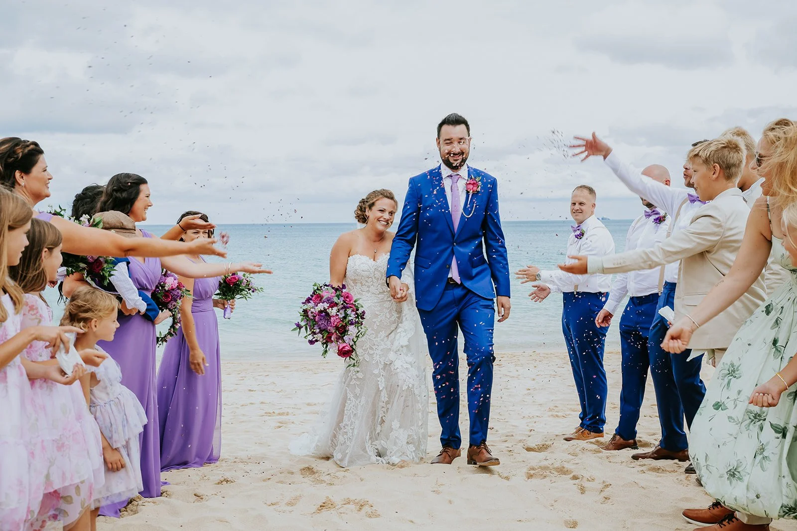 A wedding celebration on a beach with a bride and groom walking together, surrounded by friends and family throwing confetti.