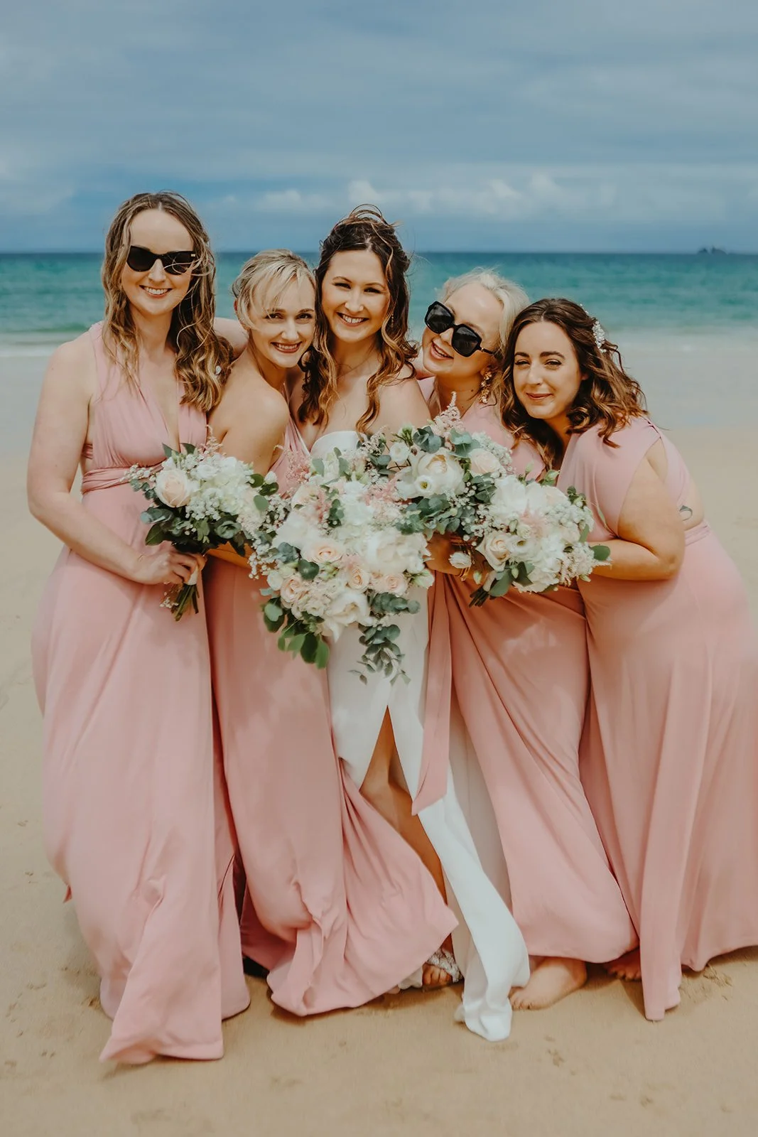 A group of five women in pink dresses standing on a sandy beach, holding bouquets, with the ocean and cloudy sky in the background.