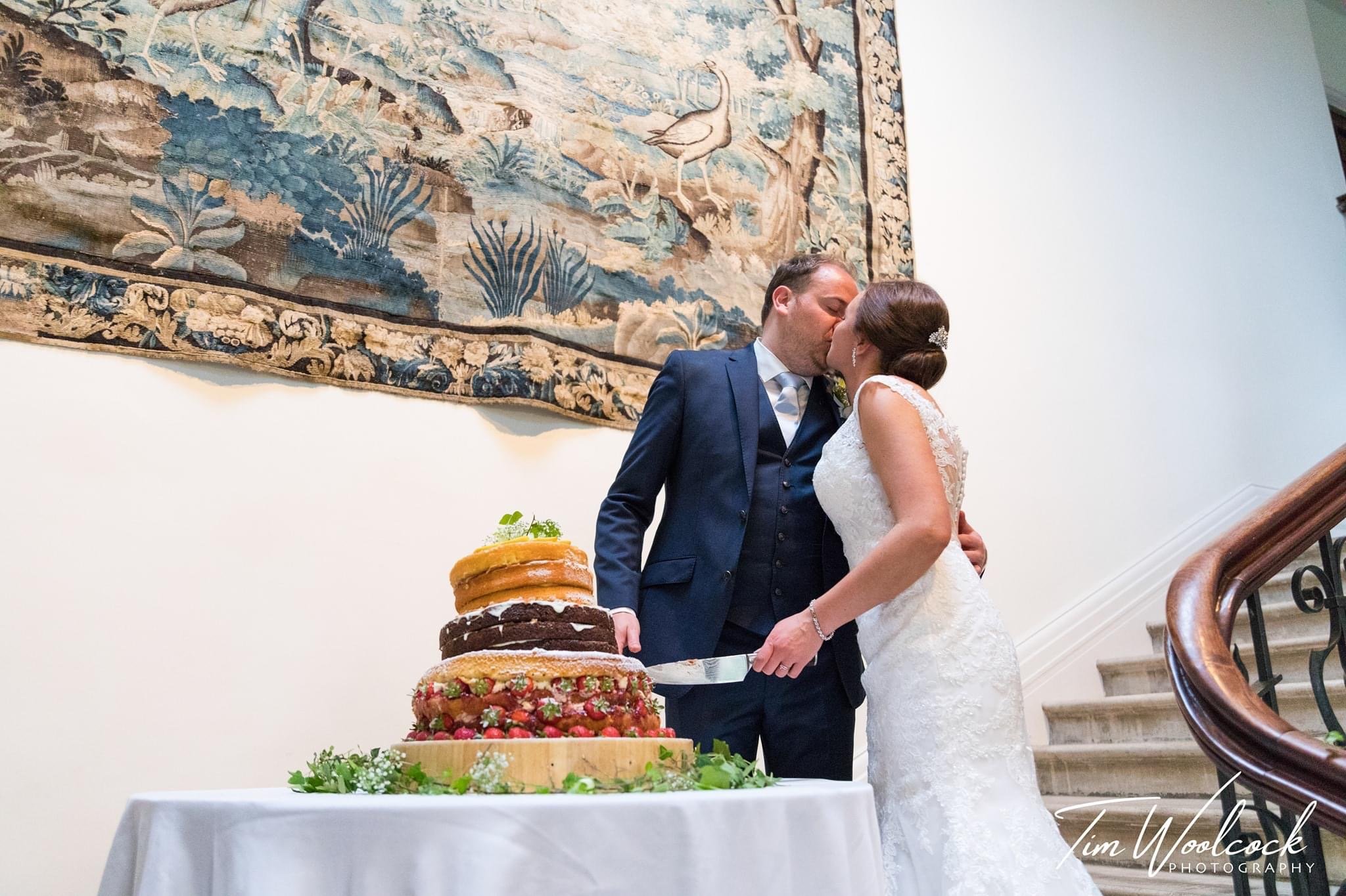 A newlywed couple shares a kiss next to a tiered wedding cake on a table, with a decorative tapestry hanging on the wall behind them and a staircase on the right side of the image.