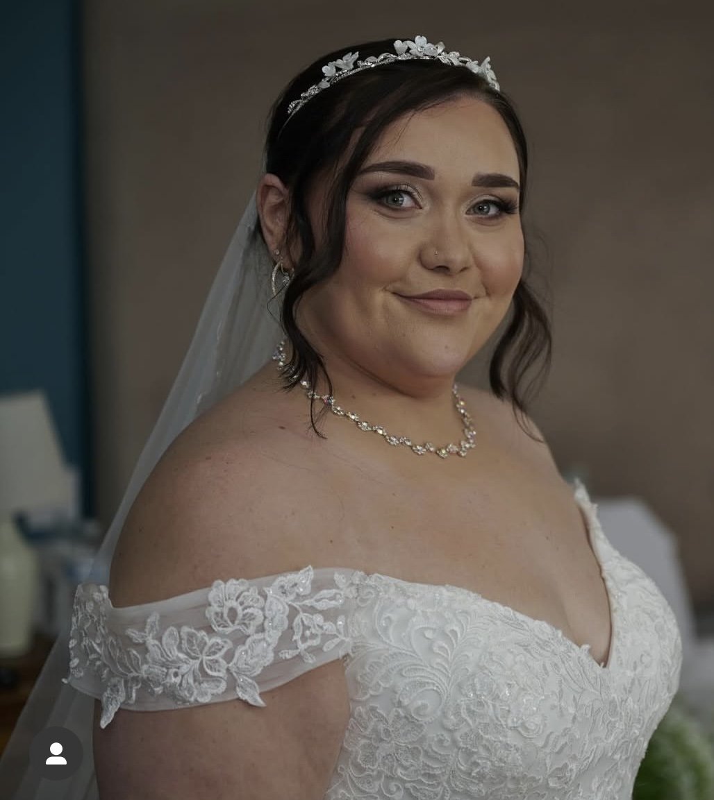 A bride with dark hair styled in loose waves, wearing a white off-the-shoulder lace wedding dress, a pearl and floral headband, a veil, and a matching pearl necklace, smiling at the camera indoors.
