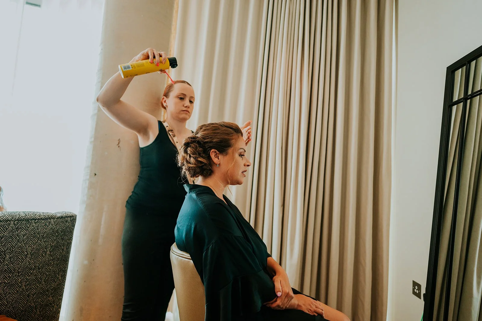 A woman with styled hair sits in front of a mirror while another woman blow-dries her hair in a room with beige curtains.