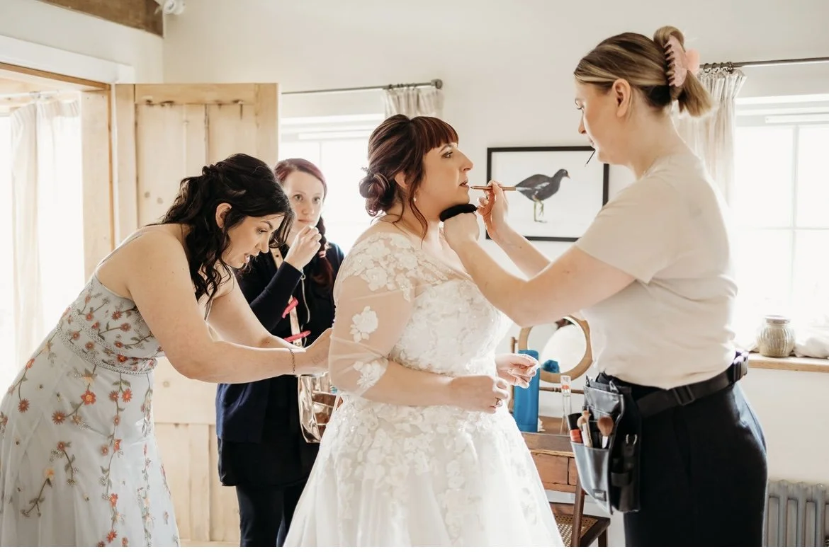 Bride getting her makeup applied by a makeup artist in a room with three other women observing. At Nancarrow farm