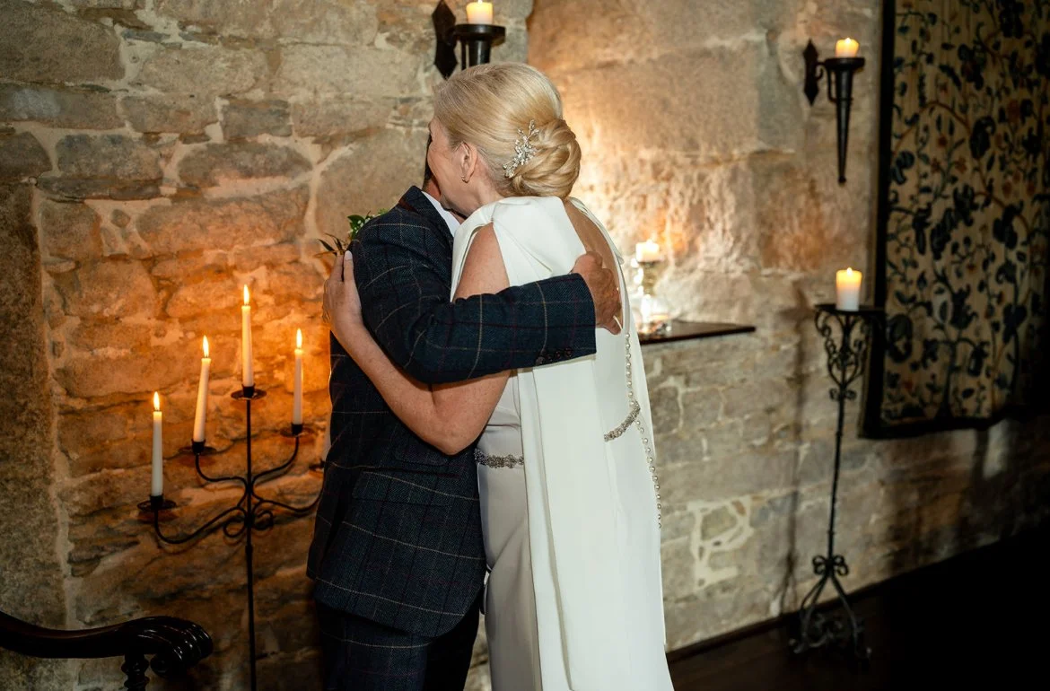 An older woman in a white dress and a man in a dark suit embrace and kiss in a warmly lit room with stone walls and candles.