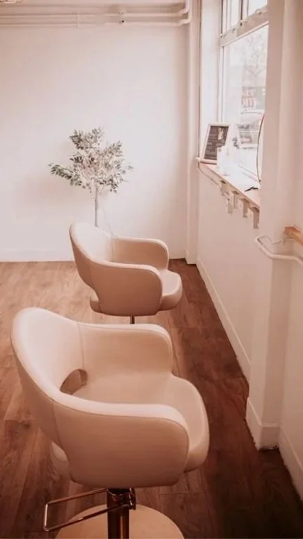 A cozy corner of a room with two beige chairs facing a window, with a small art piece and a plant on the windowsill, and light wooden flooring.