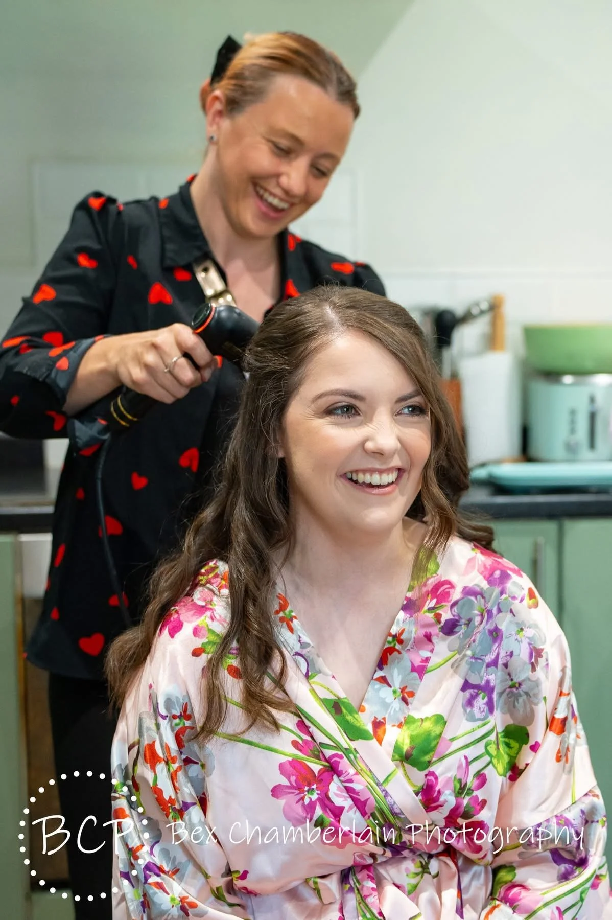 A woman is sitting and smiling while another woman styles her hair with a curling iron in a kitchen setting.