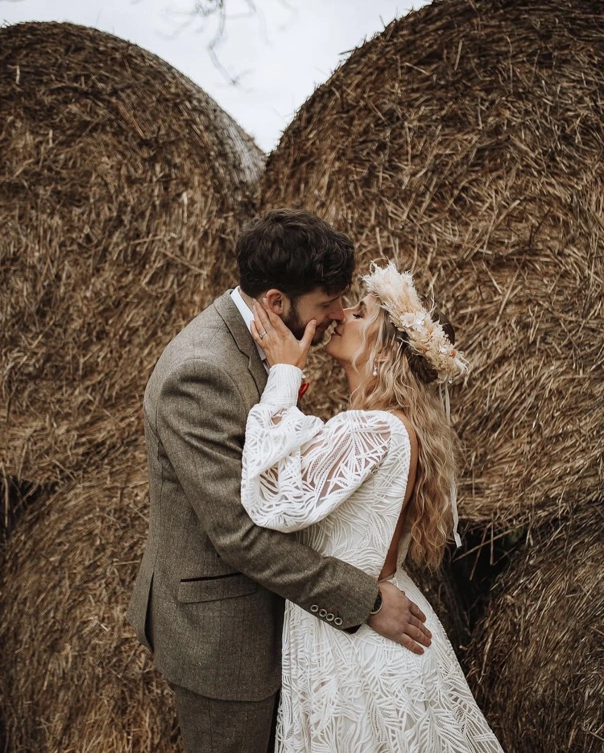 A couple dressed in wedding attire sharing a kiss in front of large hay bales, with the woman wearing a white lace dress and a wreath of flowers in her hair, and the man wearing a brown tweed suit.