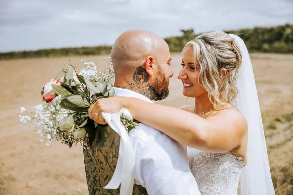 A bride and groom embrace outdoors on their wedding day, smiling at each other. The bride holds a bouquet of flowers, and the groom has tattoos on his arm and neck.