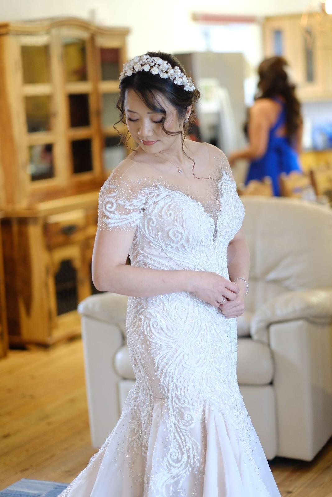 A bride in a white wedding gown with intricate embroidery and sparkles, wearing a floral headband, standing in a warmly lit room. Cottages near Barn Pengelly.