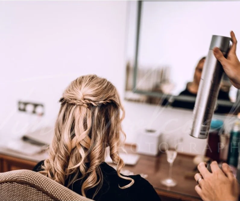Woman with blonde hair styled in loose curls and a braid at the crown sitting at a vanity mirror with a hair spray being applied.