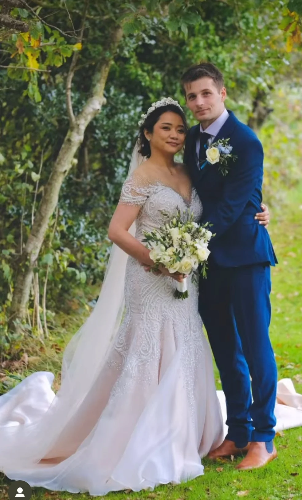 A bride and groom standing outdoors after their wedding, with green trees and grass in the background. The bride is holding a bouquet of white and green flowers, wearing a lace wedding gown with an off-the-shoulder design, and a floral headband with 