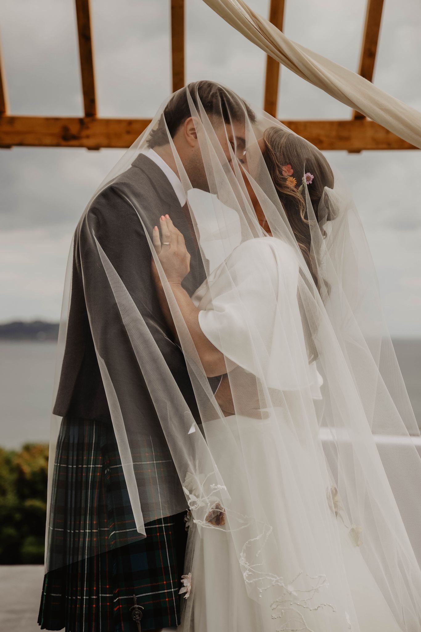 A bride and groom kissing under a wedding arch with a sheer veil covering them, outdoors by a body of water with cloudy sky. At Above the bay .