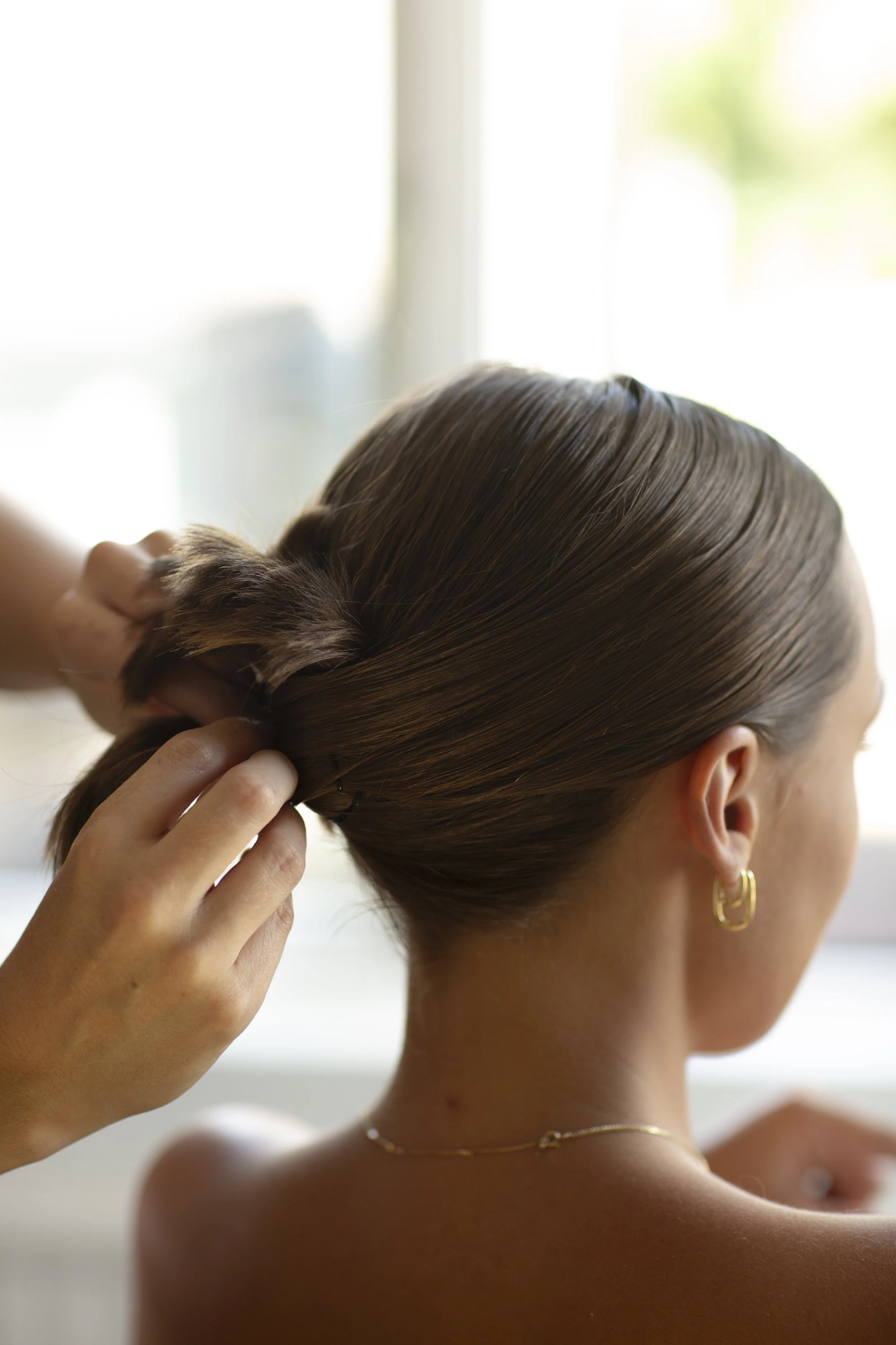 A woman with brown hair is getting her hair styled, with someone working on a low bun near the back of her head. She is wearing gold jewelry, including hoop earrings and a delicate necklace. The background shows a bright window with sunlight.
