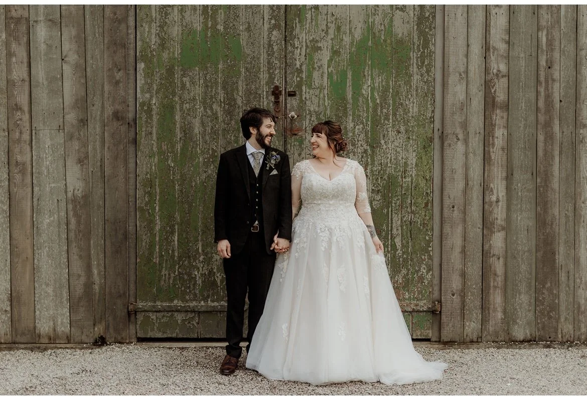 A newlywed couple holding hands and smiling at each other in front of a weathered wooden barn door.At Nancarrow farm