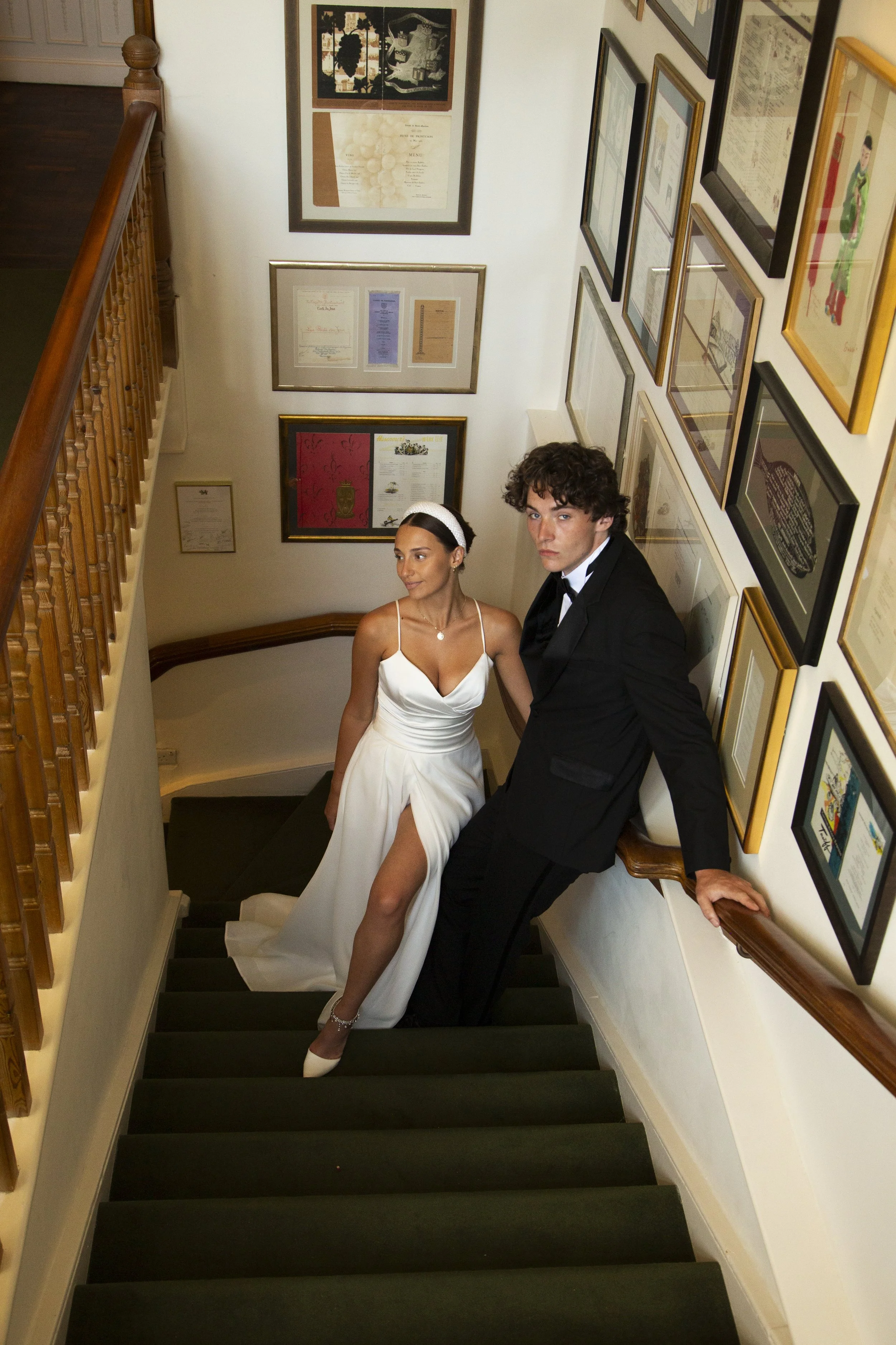 A woman in a white wedding dress and a man in a black tuxedo sitting on a staircase, surrounded by framed artwork and certificates on the walls.