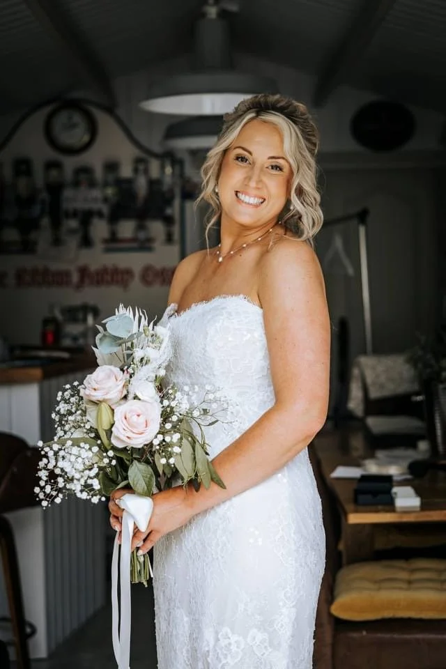 A smiling bride in a white lace wedding dress holding a bouquet of white and blush pink roses, with greenery and baby's breath, inside a rustic-style room.