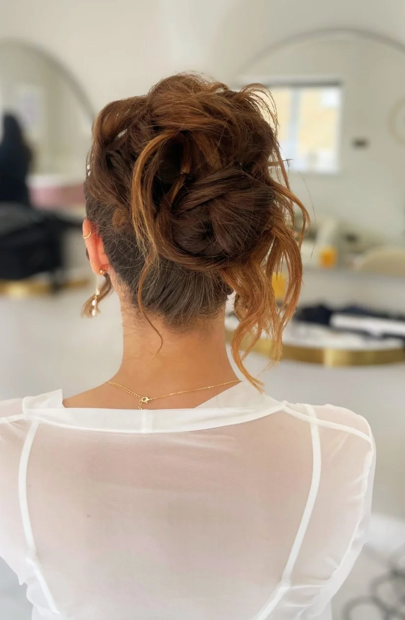 Back view of a woman with styled, curly, reddish-brown hair in an updo, wearing a sheer white top, gold necklace, and earrings, in a bright room.