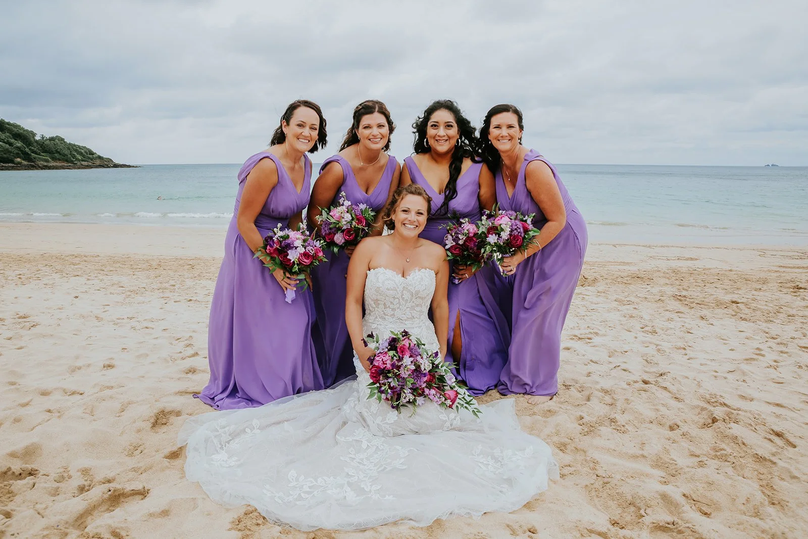 A bride in a white wedding dress sitting on the sand at the beach, surrounded by five bridesmaids in purple dresses, all holding bouquets of purple, pink, and white flowers, with ocean and cloudy sky in the background.