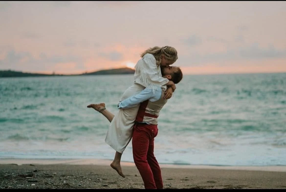 A couple is at the beach during sunset, with the man lifting the woman in the air as they embrace and smile at each other. At Lusty Glaze.
