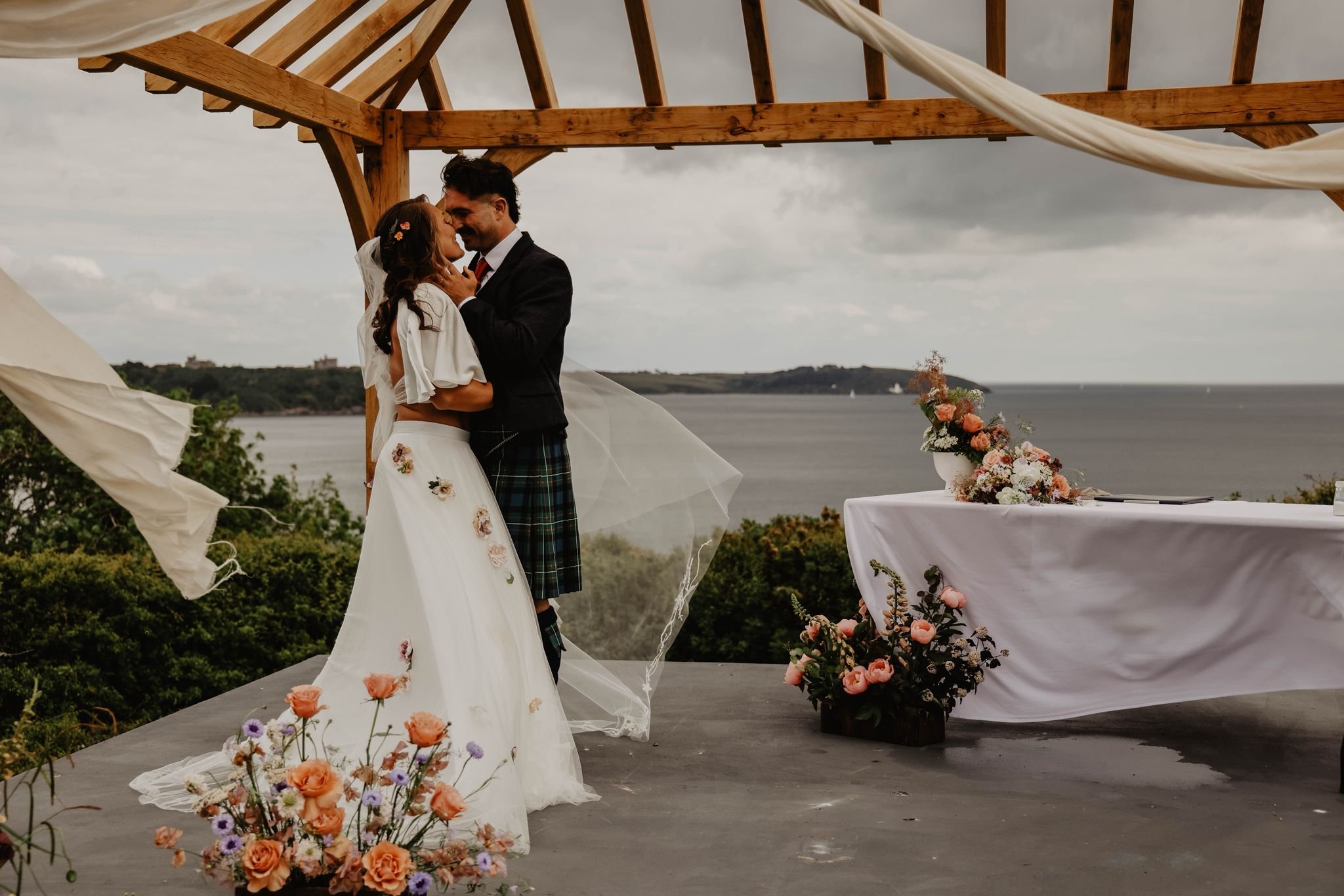 A couple getting married under a wooden wedding arch, with a scenic view of a body of water and cloudy sky in the background; the woman wears a wedding gown with floral details, and the man wears traditional Scottish attire, including a kilt.