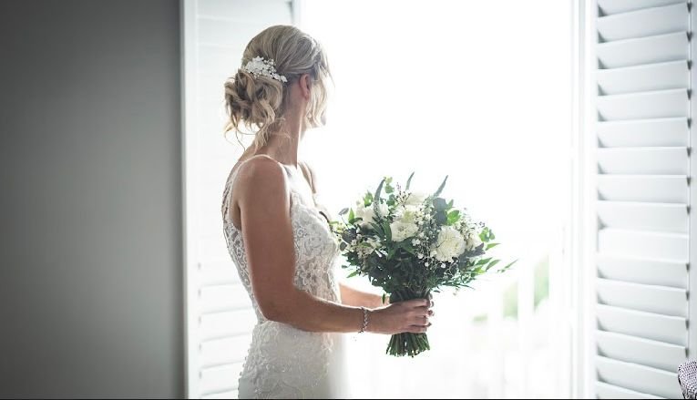 A bride in a white lace wedding dress holding a bouquet of white and green flowers, standing near a window with white shutters. At The harbor hotel St,ives