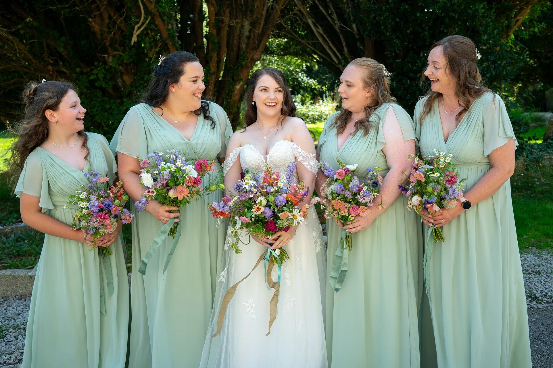 A bride and five bridesmaids standing outdoors in front of trees, all smiling and holding colorful bouquets of flowers. The bride is in the center wearing a white wedding dress, and the bridesmaids are in matching light green dresses.