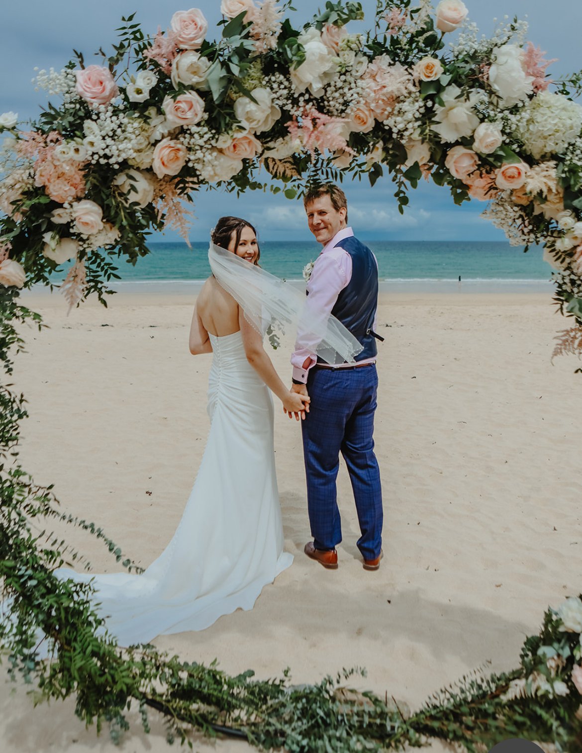 A newlywed couple holding hands on a beach under a floral arch, with the ocean in the background.