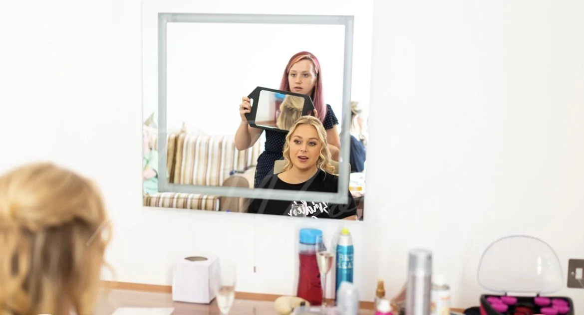 Makeup artist taking a photo of a woman with blonde hair reflected in a mirror in a salon.